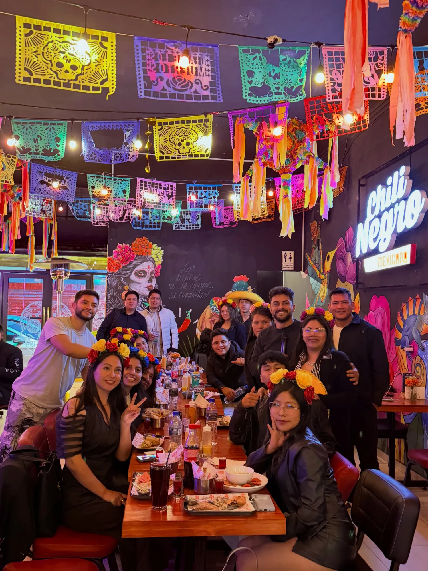 Grupo grande celebrando en una mesa larga bajo el papel picado y el letrero de neón